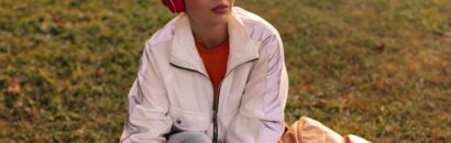 Charming short-haired woman in jeans and white jacket sits on grass. Cool girl in stylish outfit listens to music in red headphones outdoors.