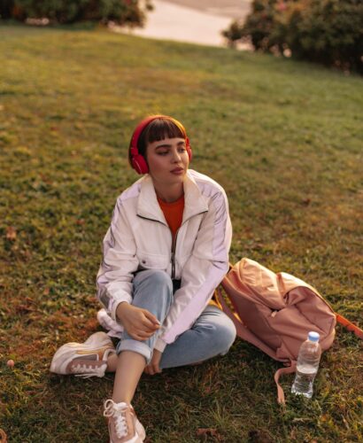 Charming short-haired woman in jeans and white jacket sits on grass. Cool girl in stylish outfit listens to music in red headphones outdoors.